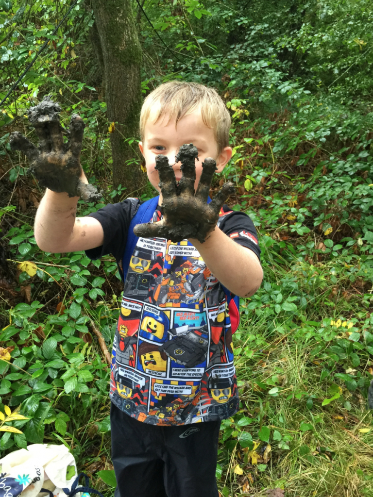 young boy holding up very muddy hands in forest