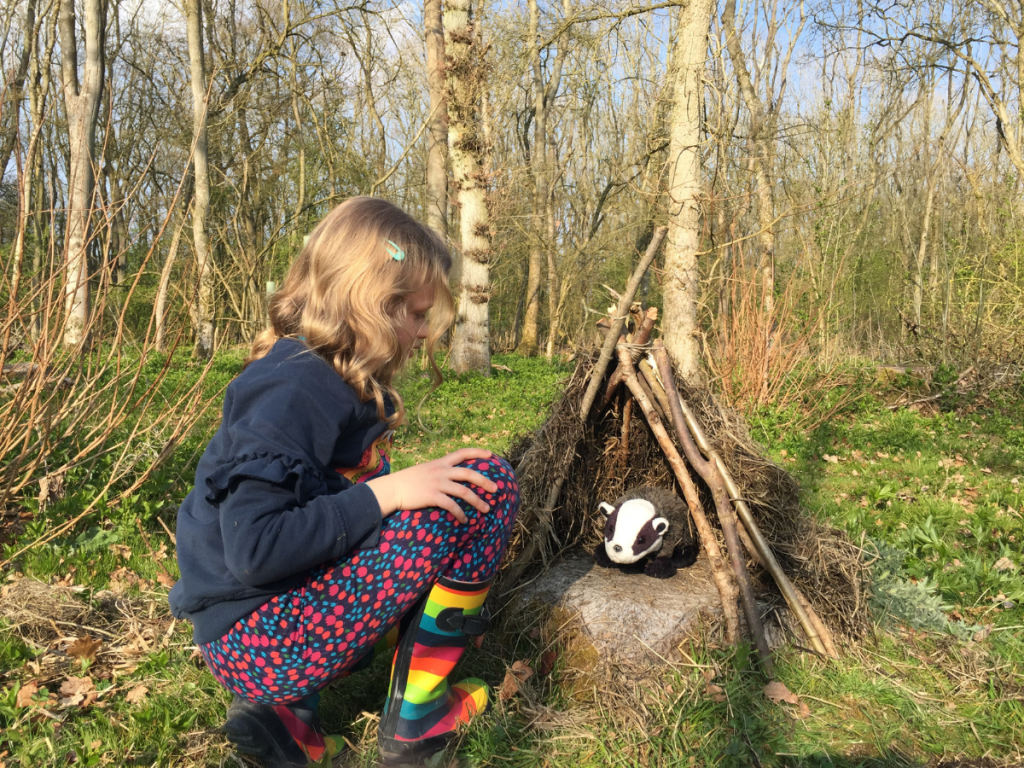 young girl in really nice stripy wellies bending over looking at a toy badger in a badger shelter in the woods.